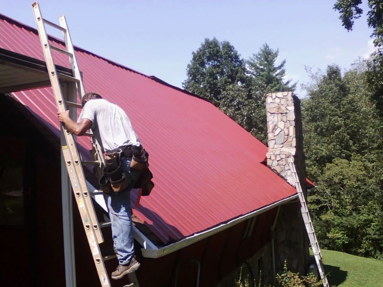 Roofer on a ladder beside a red metal roof and stone chimney — Soffit & Fascia Repair by Roofing Brothers in Dwight